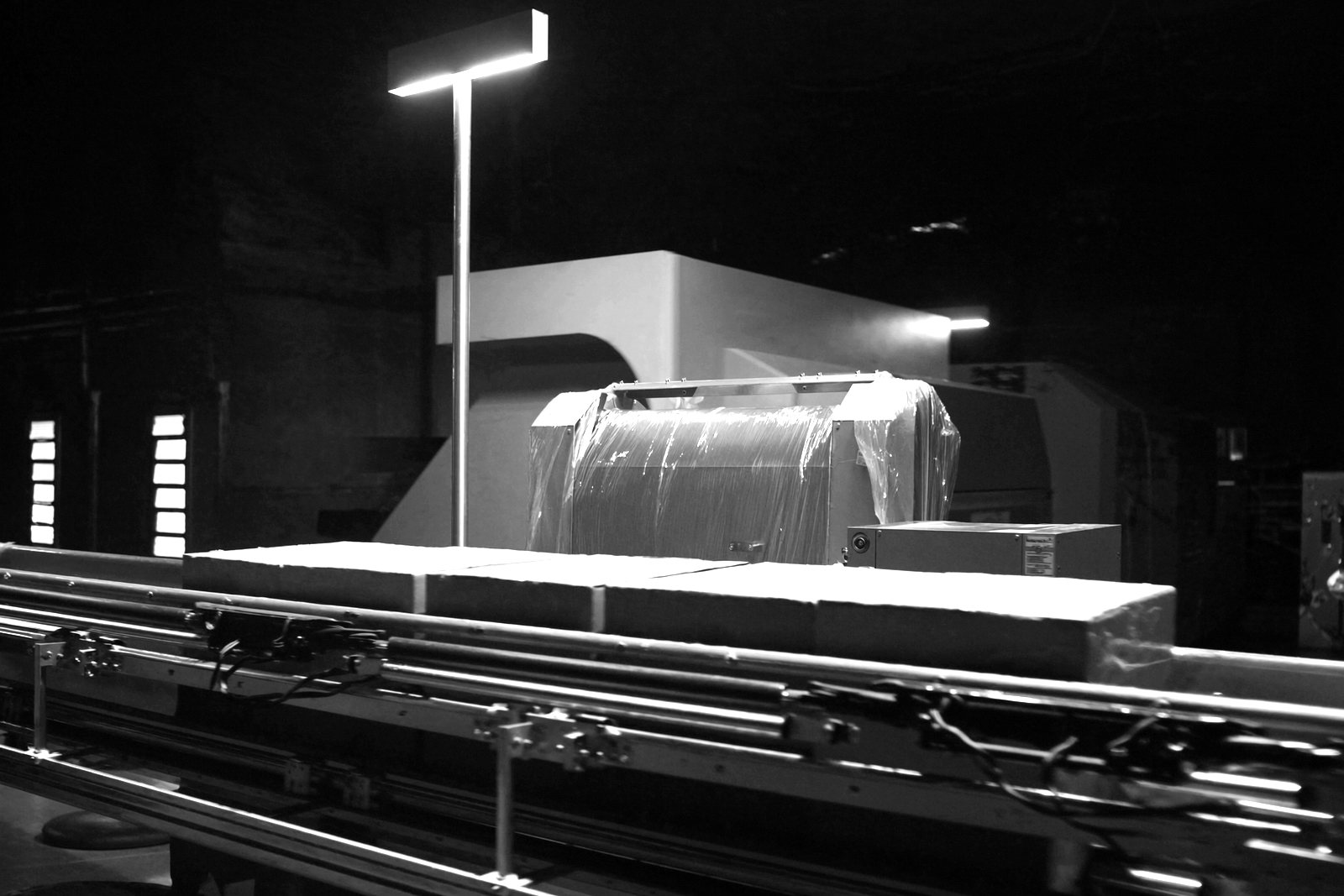 A row of sealed cardboard cases moving along a stainless conveyor past a shrink wrapper in a dark plant, lit by one overhead lamp.