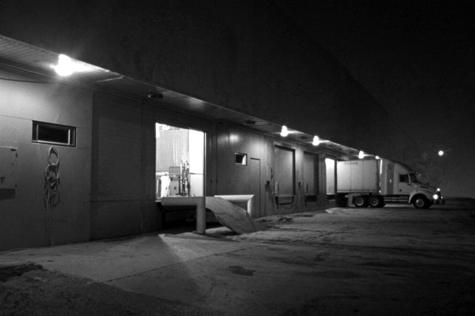 A loading dock at night under a run of lamps along the canopy, a white semi trailer backed into a far bay with its tractor still attached, a dock plate and yellow bollards at an open door in the foreground, dark sky above.