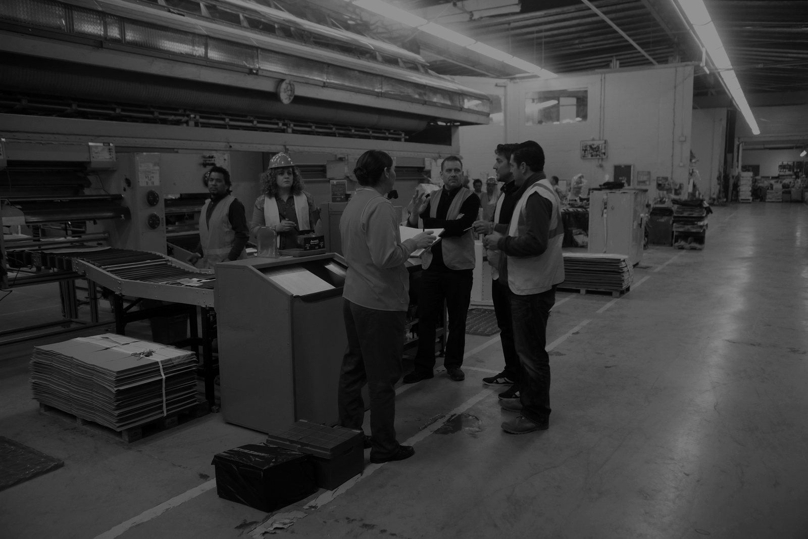Six workers in hi-vis vests standing in a loose circle beside a conveyor and a stack of flat cartons on a packaging plant floor, one of them holding a clipboard and talking to the others.