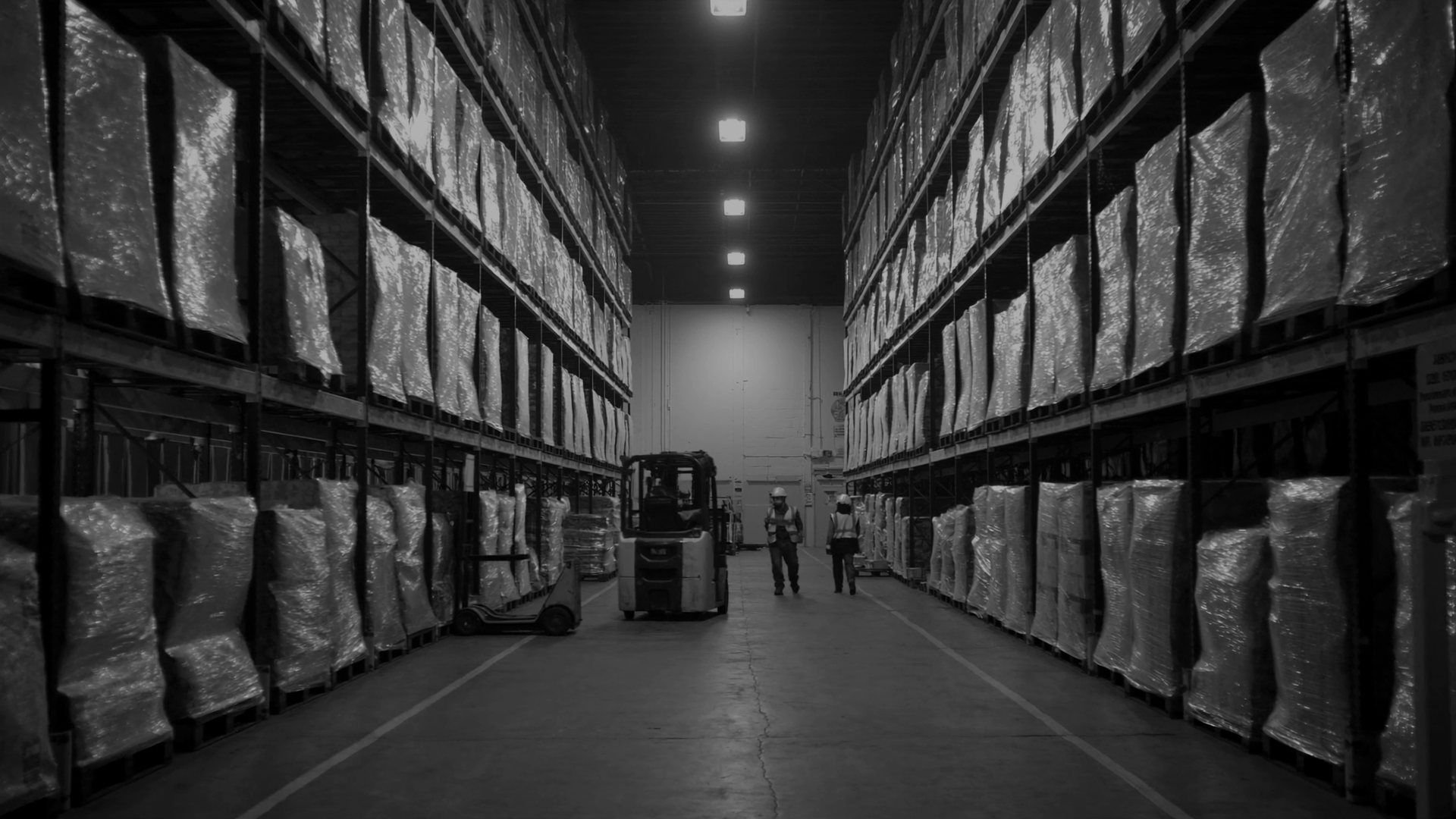 A wide aisle in a distribution warehouse at night, tall pallet racking stacked with shrink-wrapped loads on both sides, a forklift parked mid-aisle and two workers in hi-vis vests and hard hats walking toward the camera under high-bay lights.