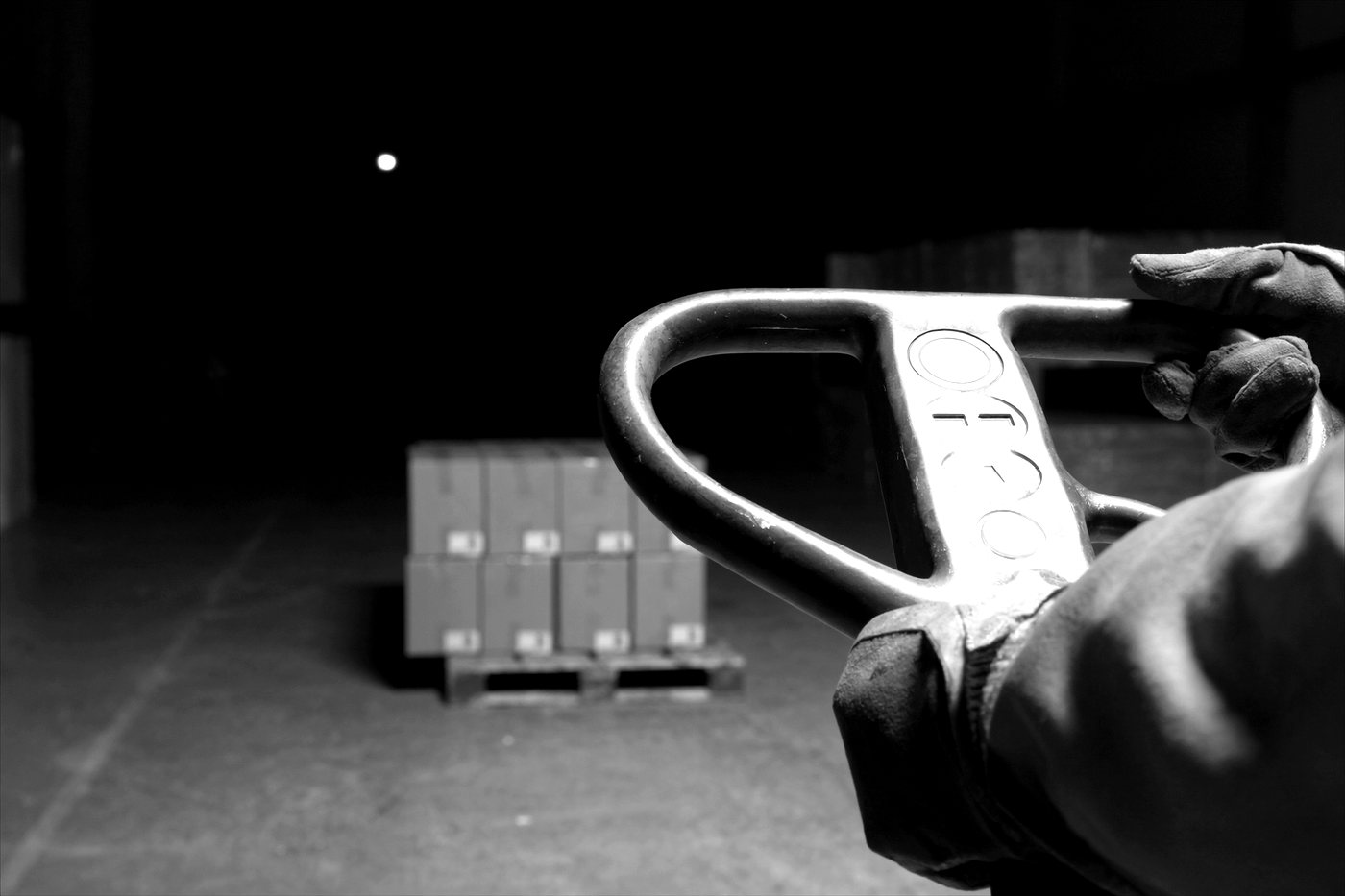 Close up of a gloved hand on the tiller head of an electric pallet jack, its control buttons catching the light, with a pallet of cartons on a dark warehouse floor behind.
